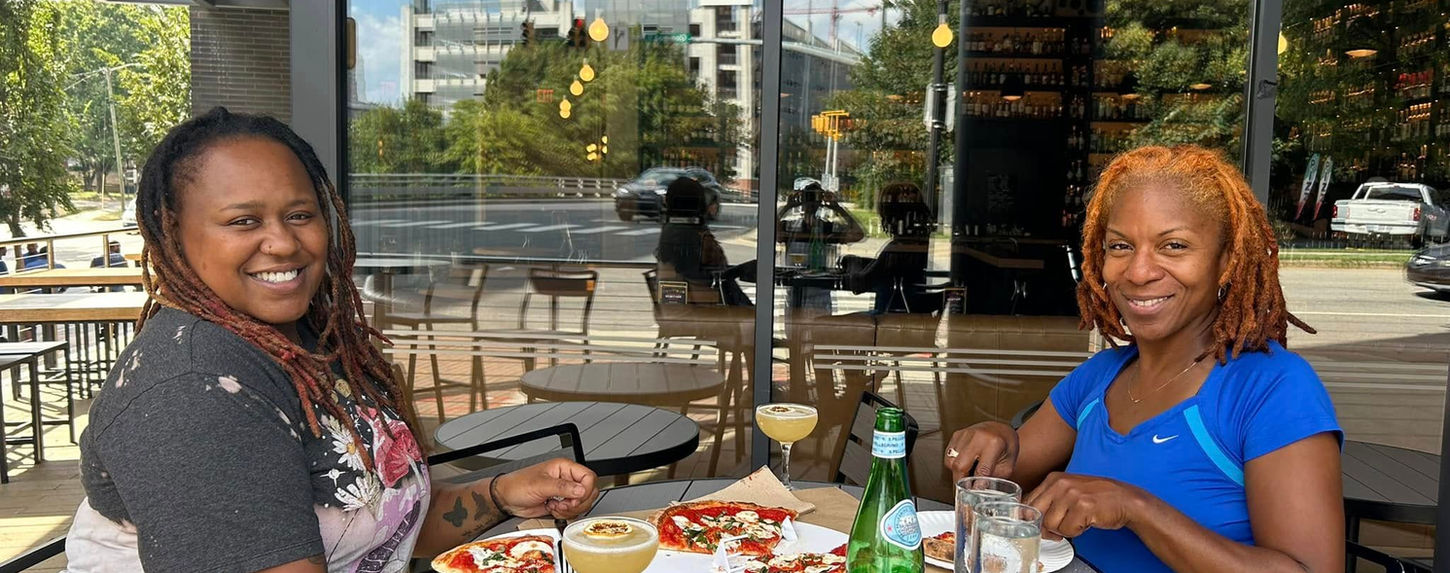 Two individuals smiling for a photo as they sit with pizza and drinks from retailers at 1 Glenwood Avenue in Downtown Raleigh