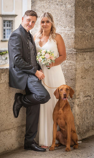 Der Bräutigam hält die Leine vom Hund und die Braut steht dahinter. Der braune Hund sitz davor.
