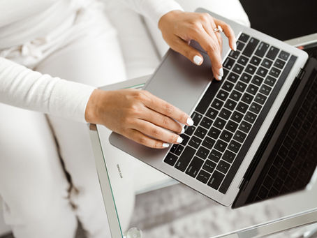 Close-up of a woman in white loungewear working on a laptop at a glass table, showcasing a clean, modern workspace and polished white nails.