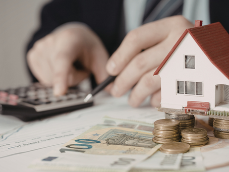 Model house on stacked coins, euro bills underneath. Hands use a calculator in the background over financial charts. Business setting.