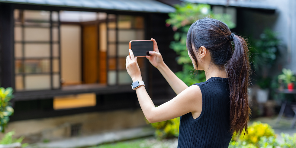Woman with ponytail photographing a modern house with her phone in a sunny garden, wearing a black sleeveless top and smartwatch.