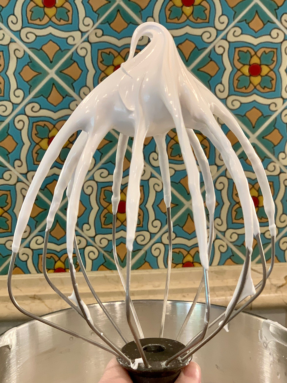 A large KitchenAid mixer whisk, being held up in front of a wall of Spanish tiles. The top has a beautiful swirl of white meringue, demonstrating the stiff peak stage.