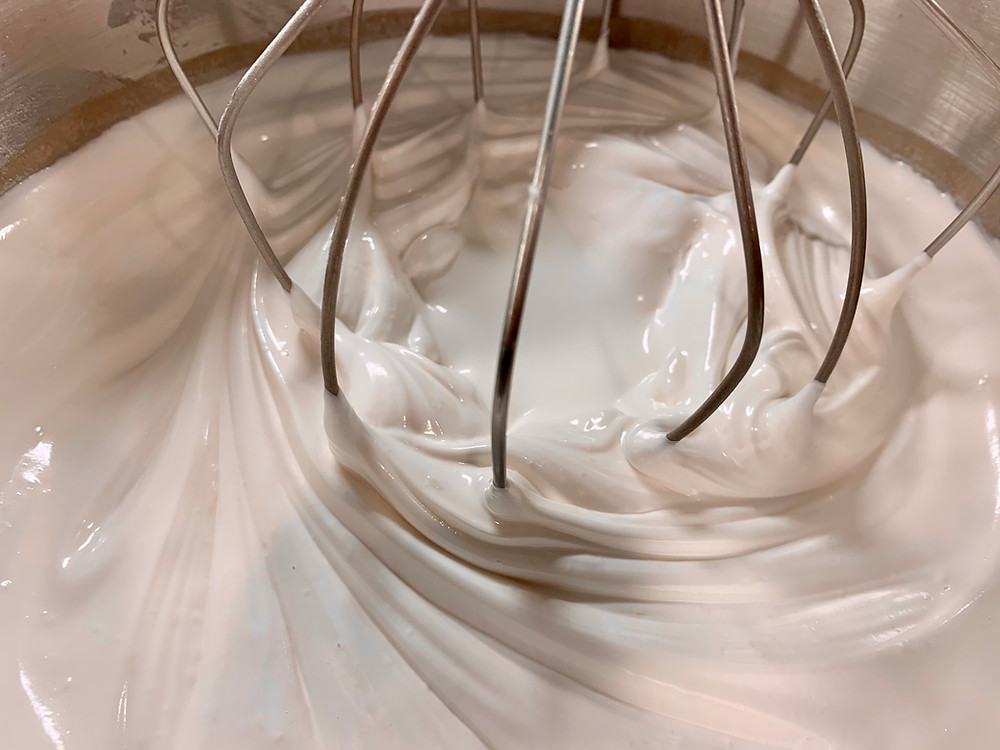 A closeup of fully whipped egg whites in a KitchenAid mixing bowl with the whisk still in it. It is a glossy and smooth white mixture that is attractively swirled around the whisk.