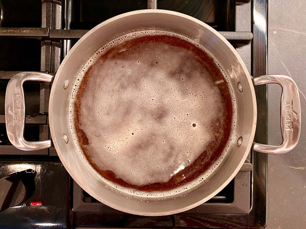 Foaming dark colored apple cider syrup in a small, shallow pot on the stove.