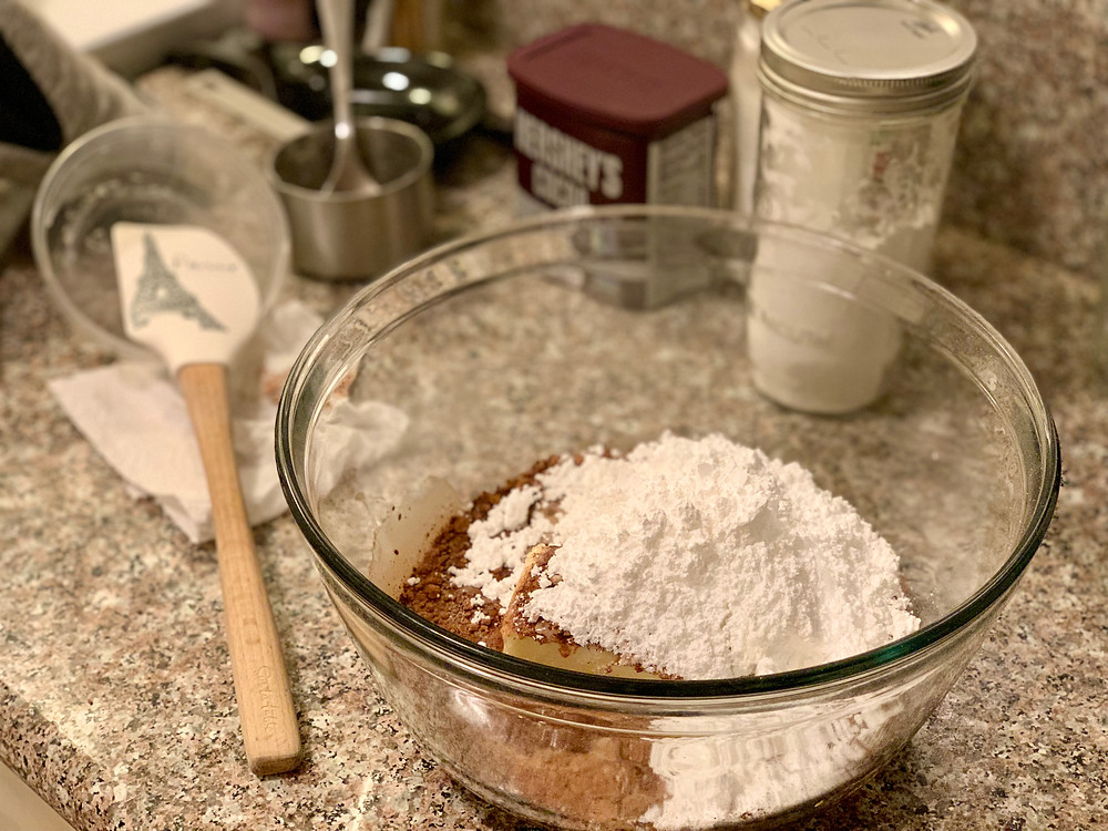 All of the buttercream ingredients in a clear mixing bowl/ the powdered sugar is on top. There is a wooden and silicon spatula printed with the Eiffel Tower to the left of the bowl.