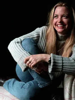 A photo of lawyer, writer, and forever New Yorker Elizabeth Wurtzel, a pretty, lively blonde woman in early middle age. She is wearing a beige sweater and jeans, and the photographer has captured her in a relaxed, smiling moment when her hands are clasped upon one knee. 
