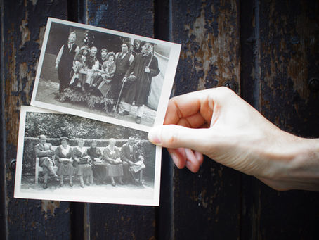 Hand holding two vintage photos of groups of people in old-fashioned clothing, indicating previous generations. Background is a rustic, wooden surface. No text visible.