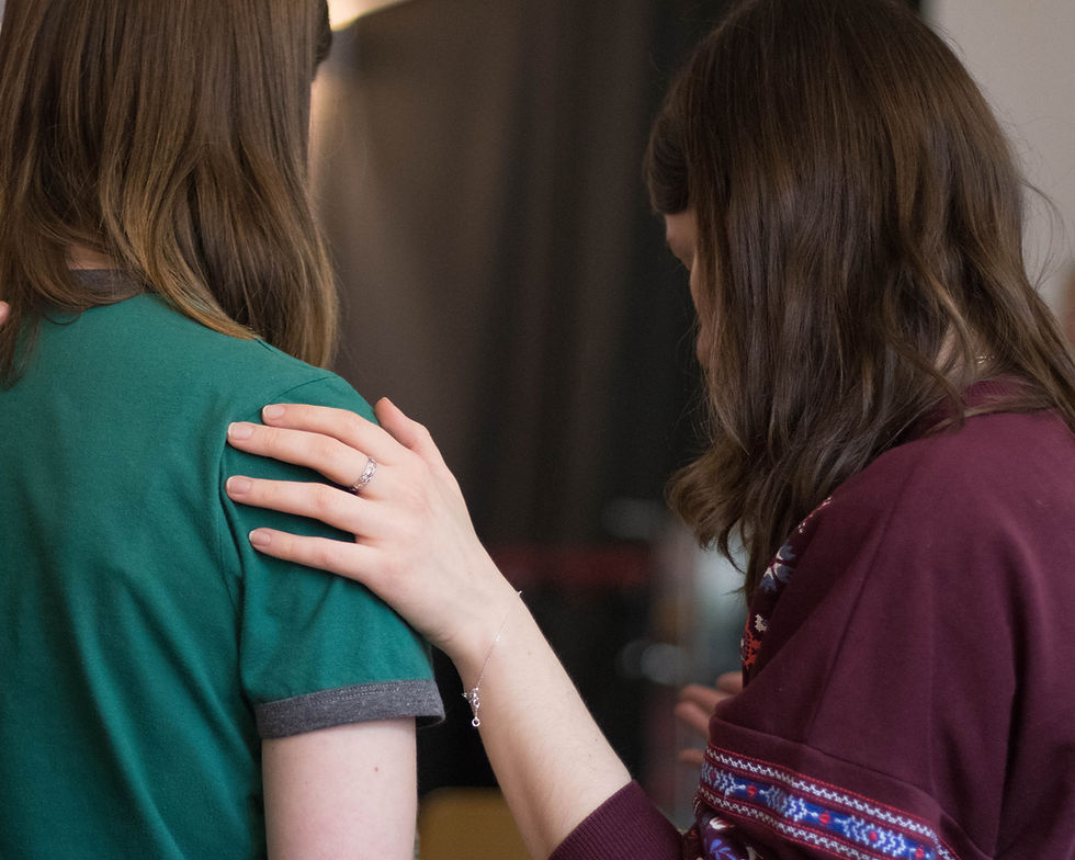 Two people with brown hair are shown from behind. One gently rests her hand on the other's shoulder. The setting is indoors, with a calm mood.