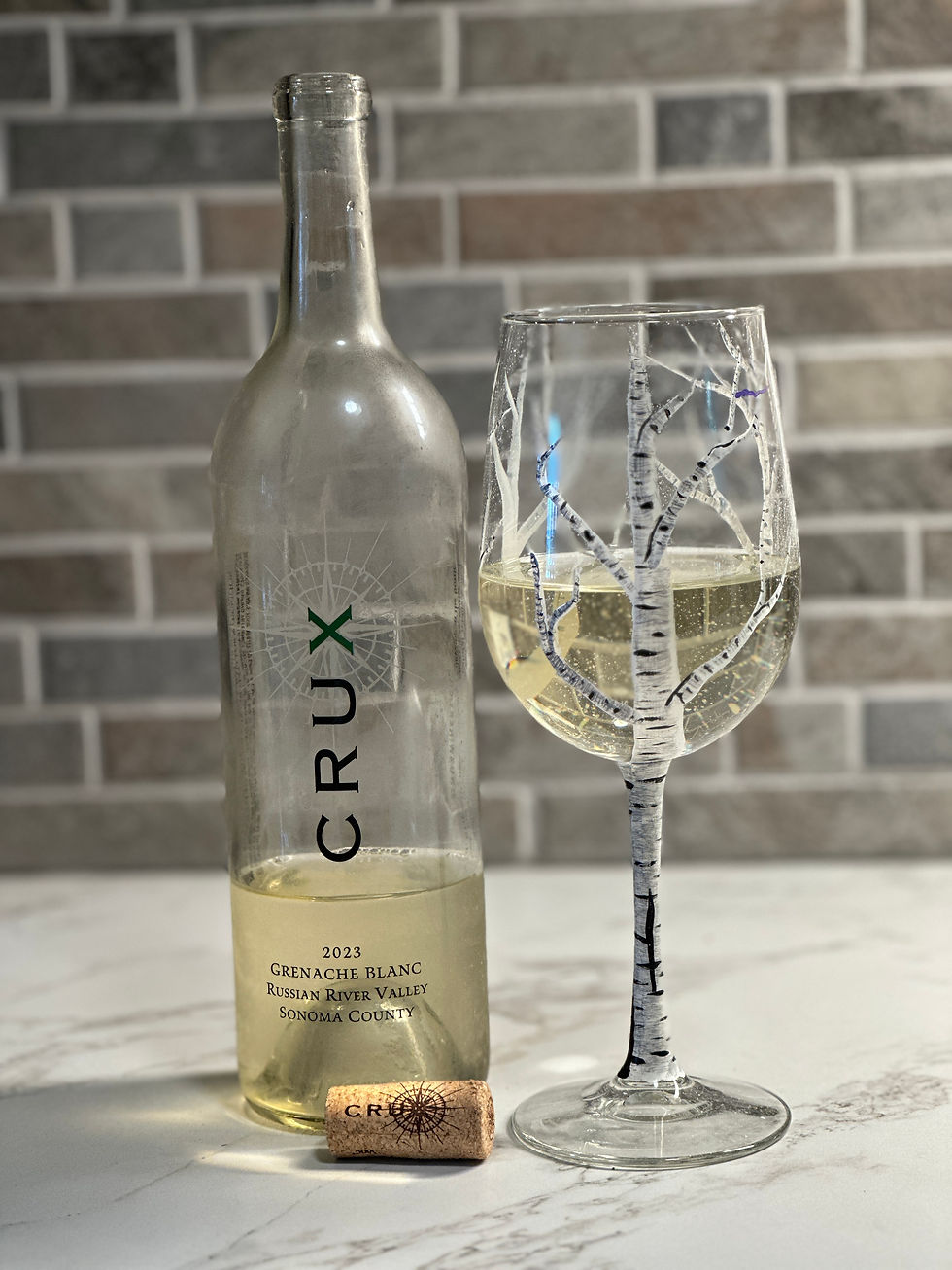 Wine bottle and glass with white wine on marble. Bottle reads "CRUX" and "2023 Grenache Blanc." Cork nearby. Brick wall backdrop.