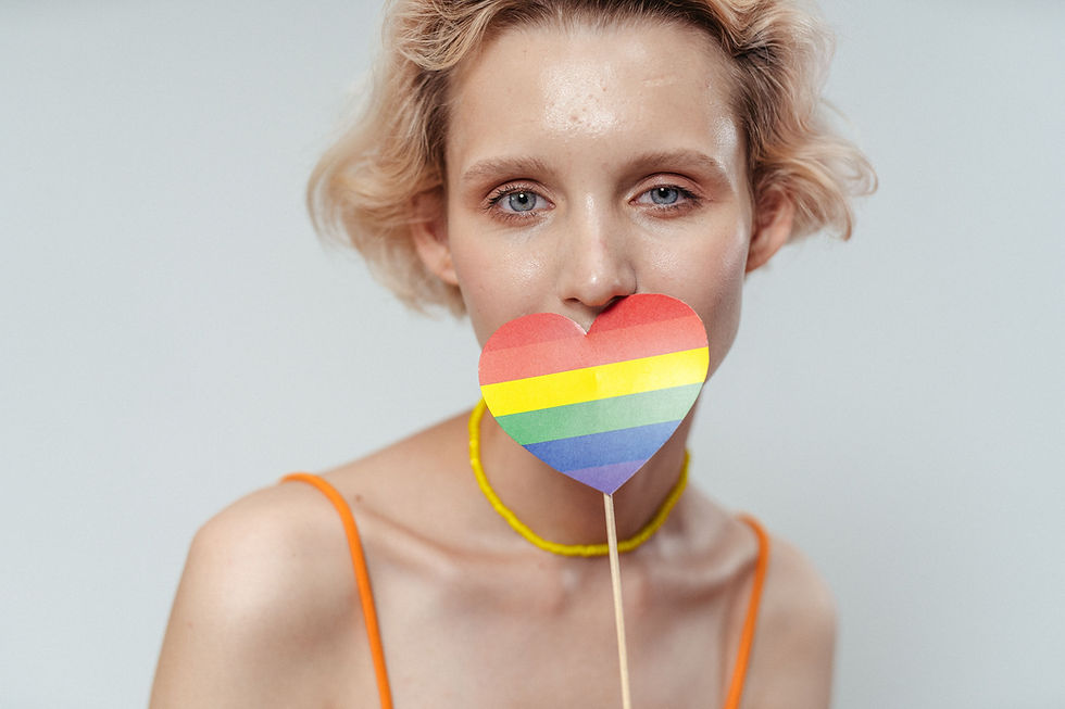 Portrait of a blonde woman holding a rainbow heart lollipop over her mouth, with a plain light background and calm gaze.