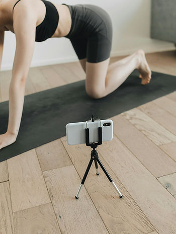 A woman practices yoga on a mat, with a phone mounted on a tripod beside her for recording or streaming.
