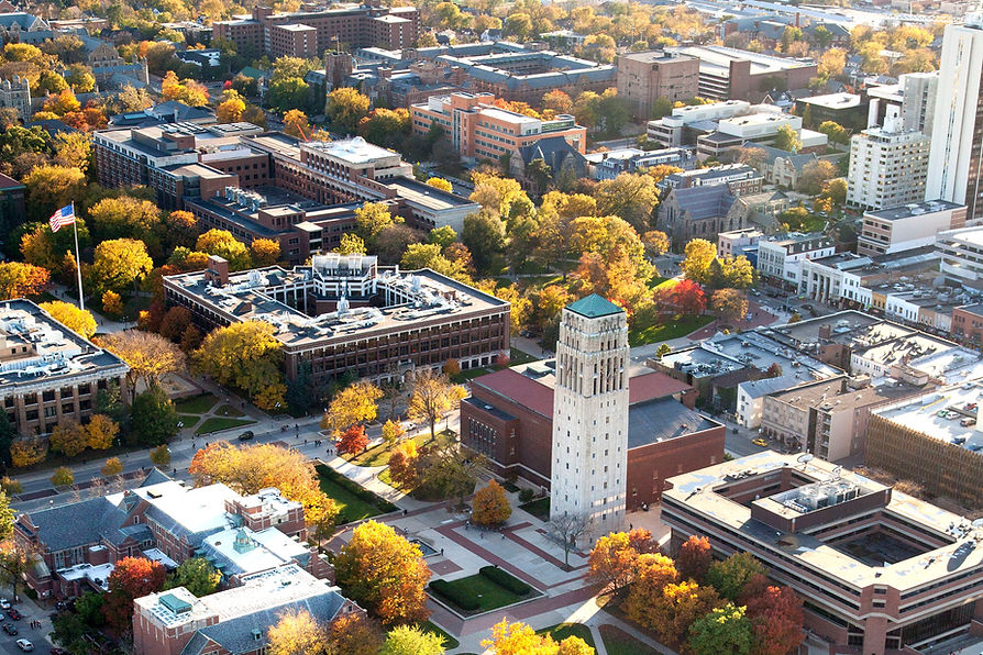An overhead view of the University of Michigan campus. Part of the diag to the top left, Ingalls Mall and the clock tower to the bottom, and a section of State Street to the right. There are brick buildings of mostly red-brown brick throughout. The center of the diag holds a waving American flag. The clock tower is a stark white grey marble against the red buildings around it. 