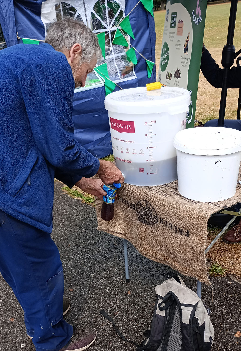 Older man in blue jacket pours liquid from a bottle into a white bucket at an outdoor stall with recycling signs.