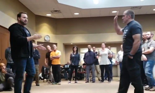 Two men demonstrate martial arts moves in a room with a group of onlookers. The setting is bright and casual, with people observing intently.