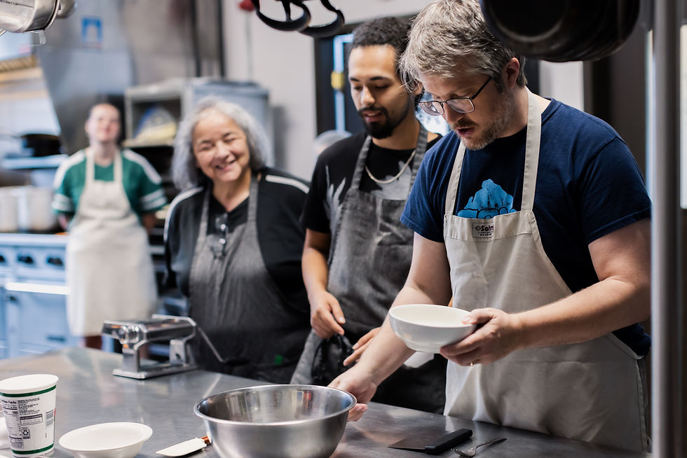 A man in white apron holding a white bowl in a kitchen while two people observe and smile