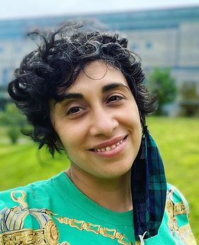 A photo of Larissa smiling at the camera, a mask hangs off their left ear.