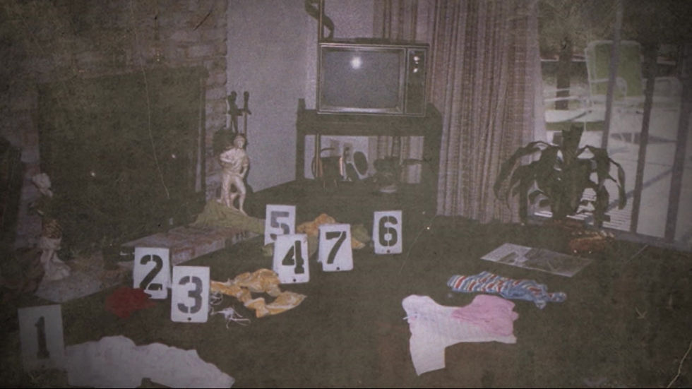 A grainy, vintage crime scene photograph of a living room with green carpet. White evidence markers numbered 1 through 7 are placed on the floor next to scattered pieces of clothing. The room features a brick fireplace on the left, an old CRT television set on a stand in the center, and a sliding glass door with curtains on the right.