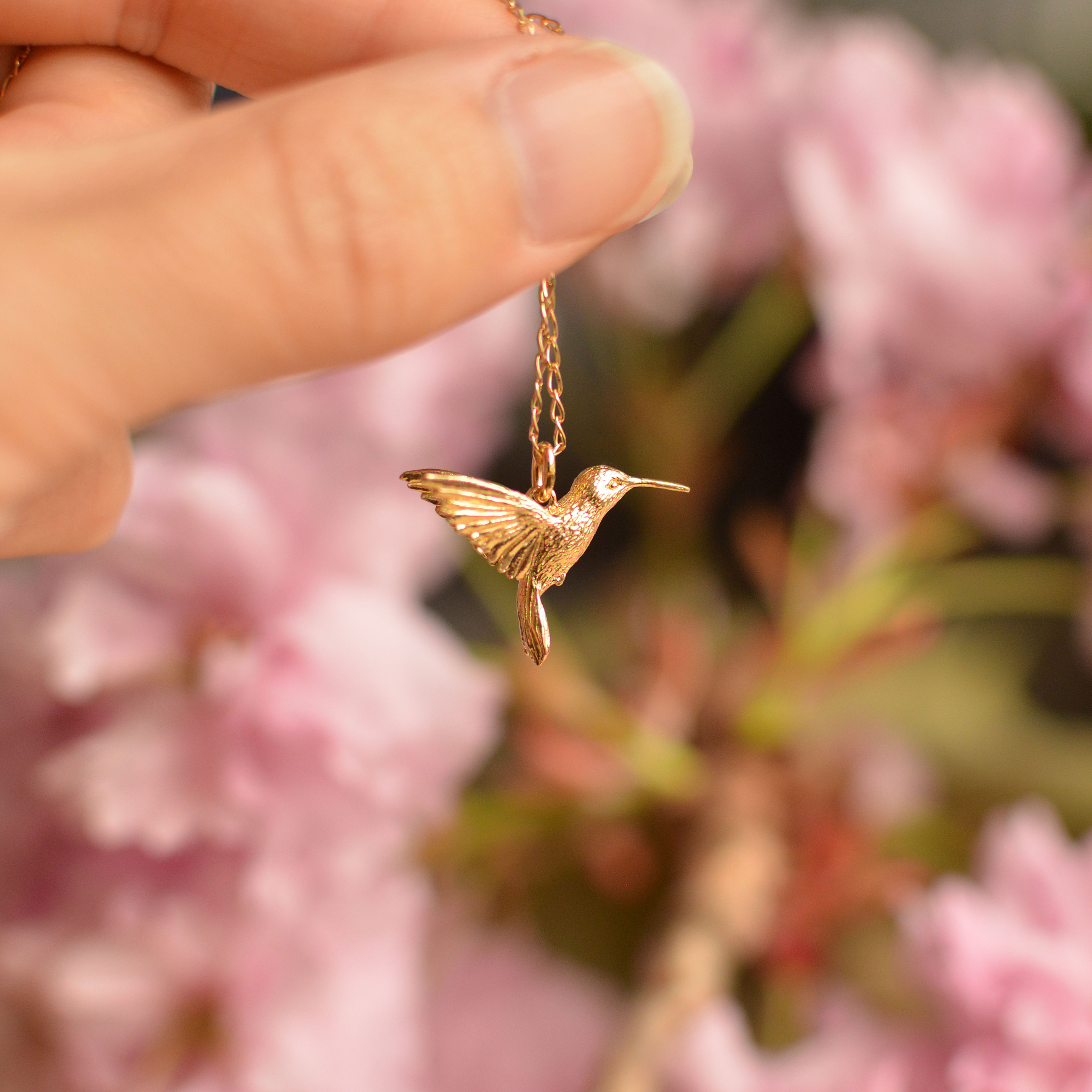Gold Hummingbird pendant on chain held against blossoms