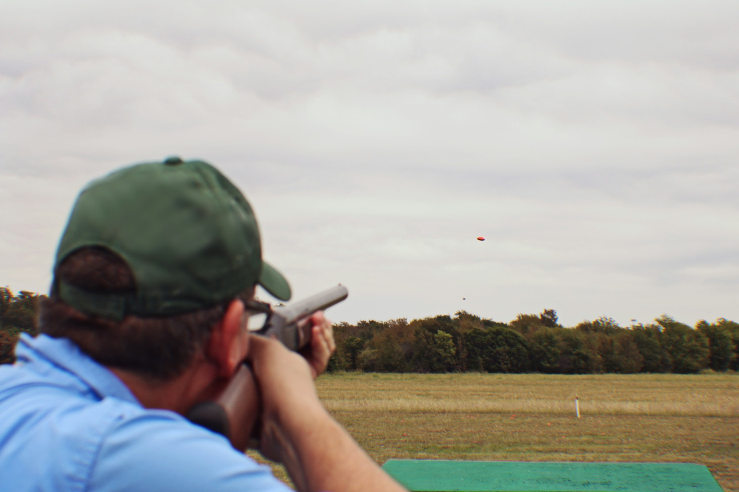 A shooter in a green cap, seen from behind, aiming at an orange clay target over an open field