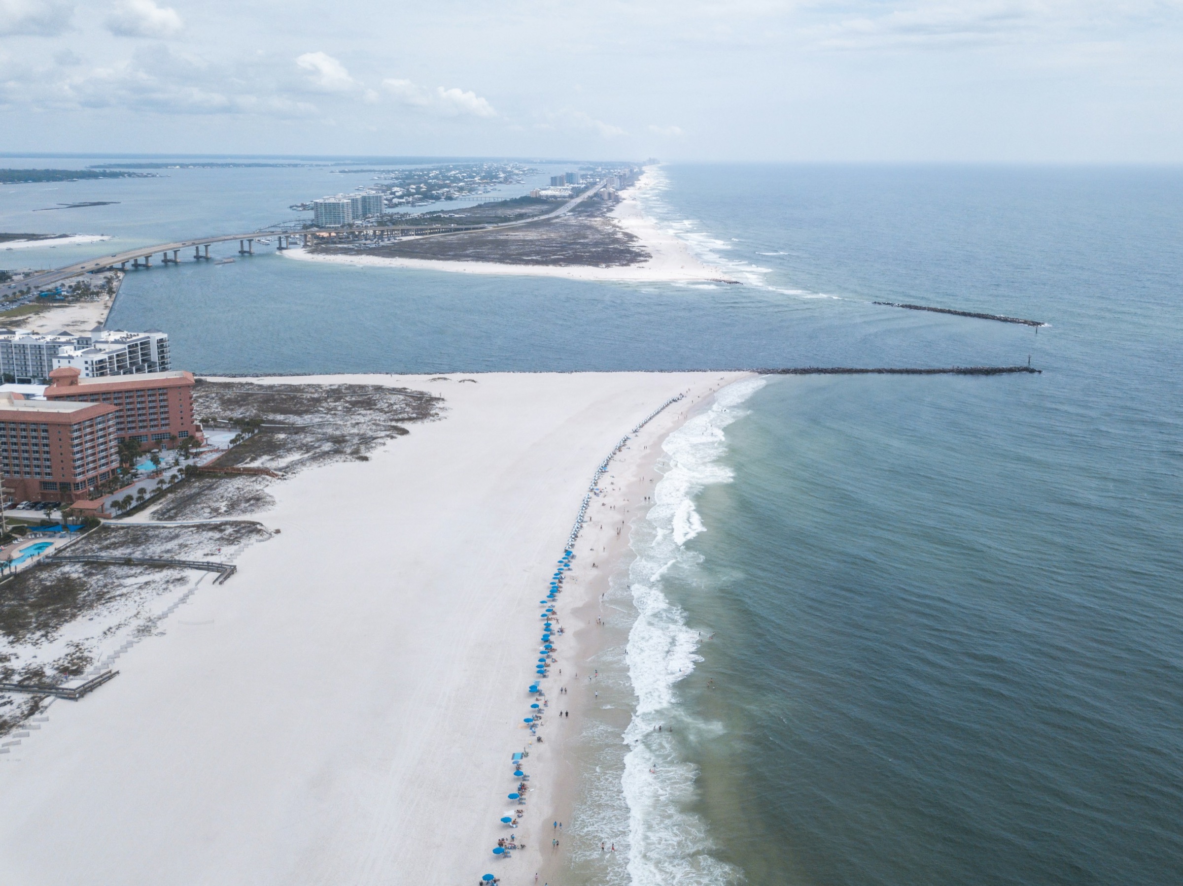 An aerial view of a white-sand Gulf beach, an inlet pass and a jetty with surf rolling ashore