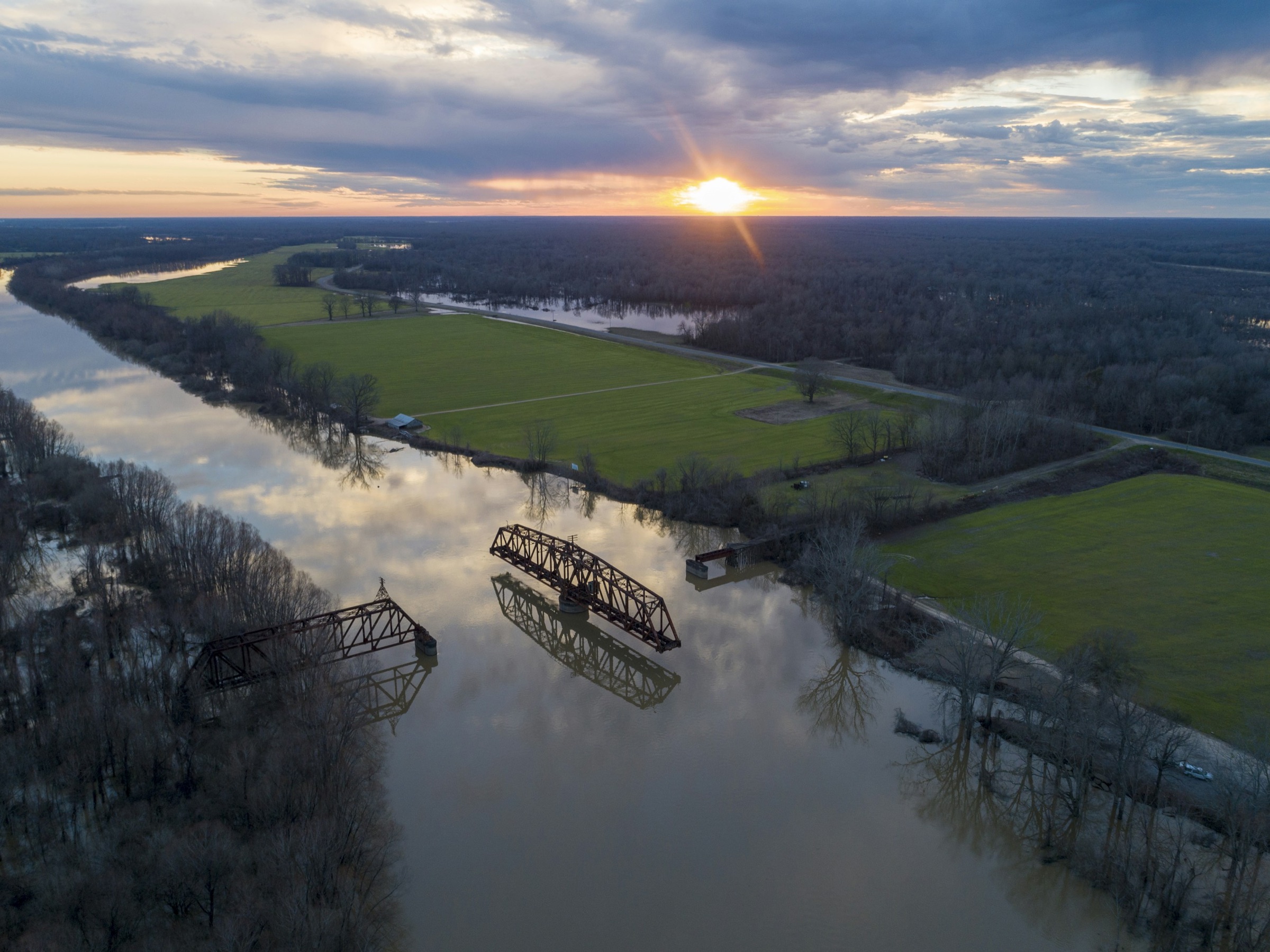 An aerial view of a river winding through flooded bottomland forest and green fields at sunset