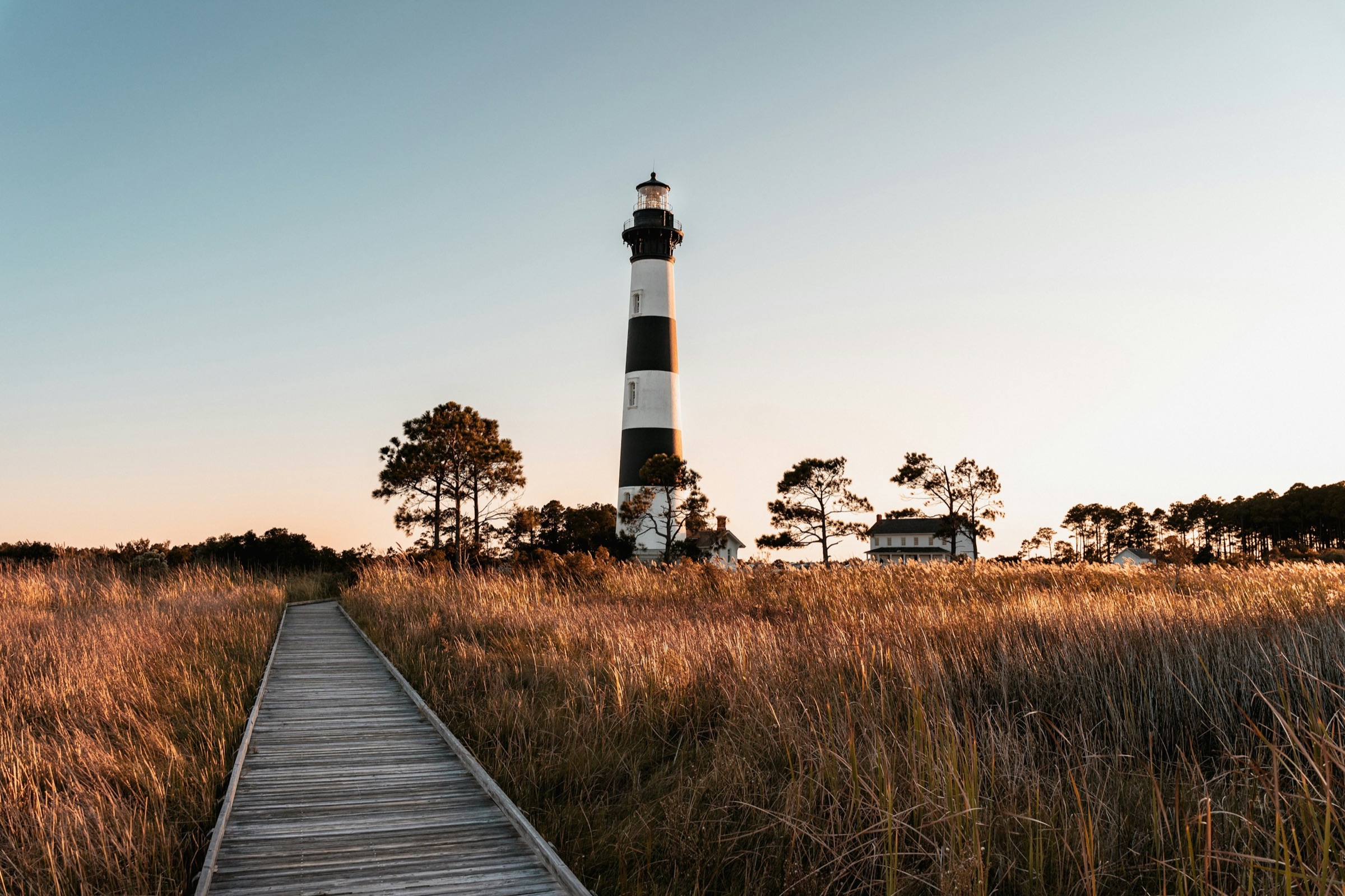 A black-and-white striped lighthouse rising above golden marsh grass, with a wooden boardwalk leading toward it