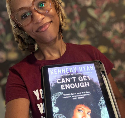 A black woman has her blond locs styled in a bob and wearng a maroon shirt with white letters and glasses with big lenses and pink frames and is holding an iPad with a book image in front of her.