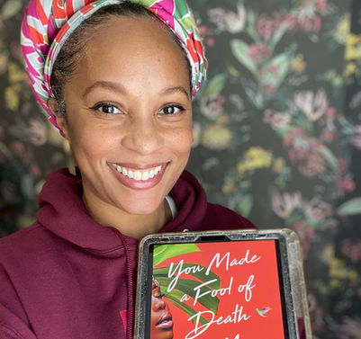 A smiling black woman, who's hair is in a colored head wrap, is holding up a book.
