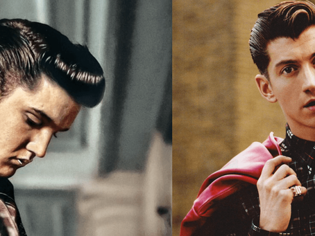 Two men with slicked-back hair; left wears a patterned suit looking down, right wears plaid with red tie holding a pink jacket, urban setting.