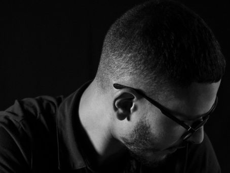 Man in glasses looks down against dark background, wearing a dark shirt. The image is monochrome, creating a contemplative mood.