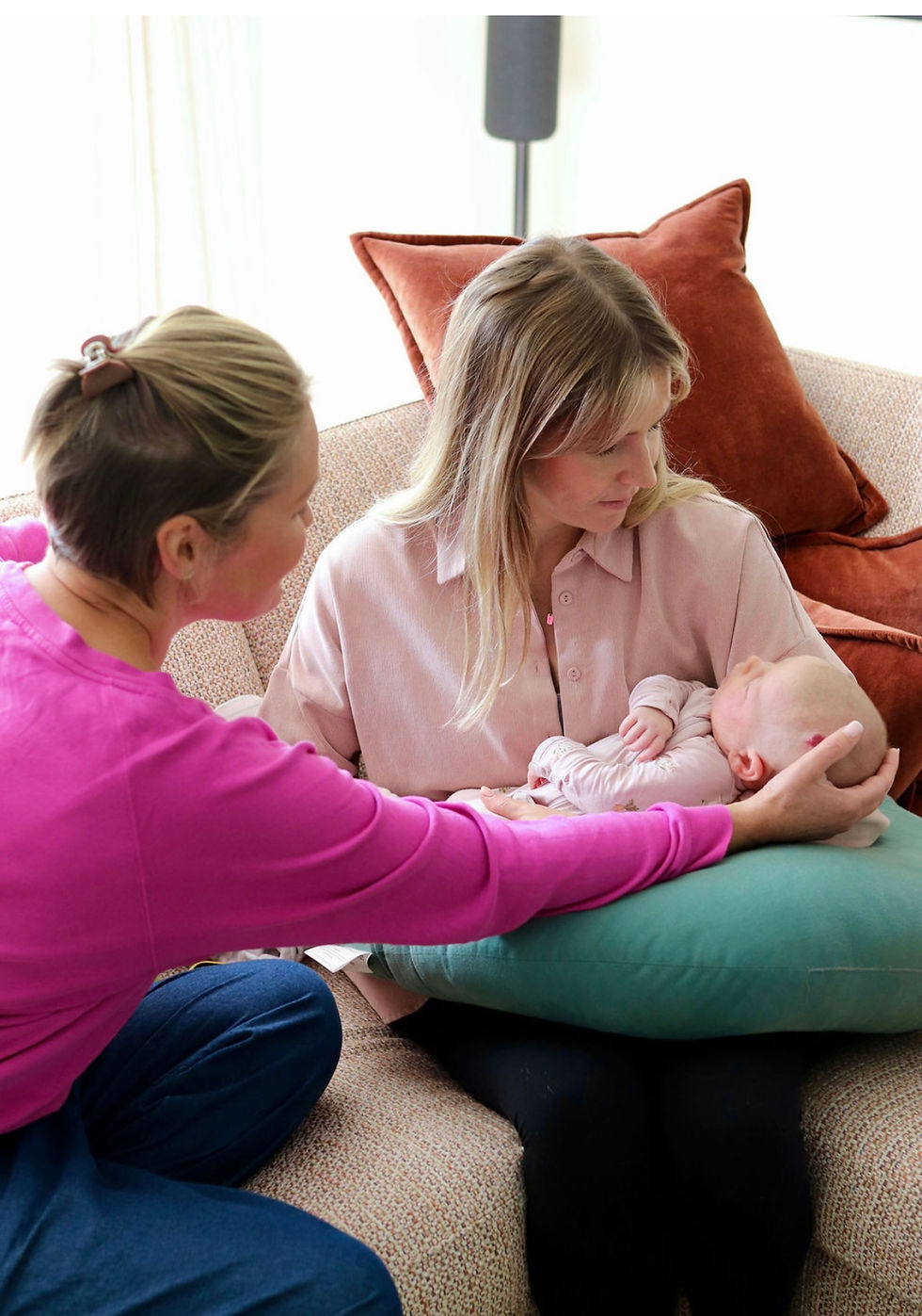 Two women on a couch gently cradle a sleeping baby, with rust and teal pillows in a bright living room, calm and tender.