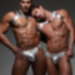 Two men pose in metallic harnesses and briefs. One rests his head on the other's shoulder. The background is neutral and dimly lit.