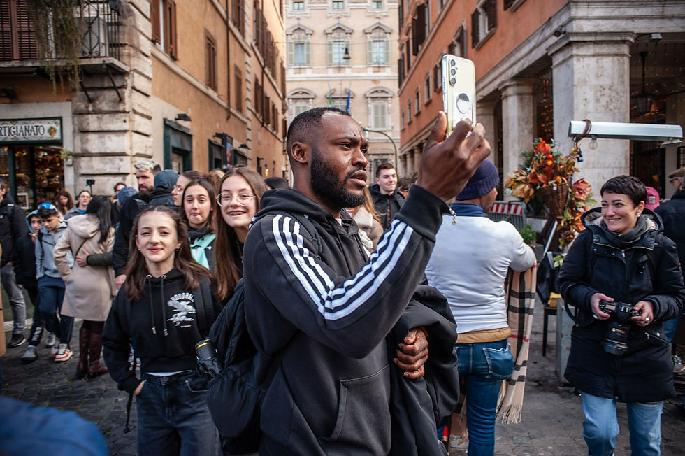 Turisti e pellegrini nel centro storico di Roma durante il Giubileo 2025. Un uomo con felpa sportiva nera scatta un selfie tra la folla, mentre gruppi di giovani sorridono e una fotografa impugna la sua macchina fotografica. Le strade affollate e le decorazioni natalizie creano un’atmosfera vibrante tra i vicoli della città eterna.