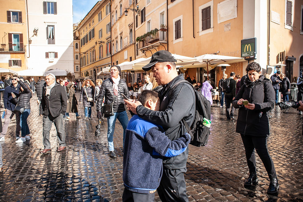 Turisti e pellegrini nella piazza del Pantheon a Roma durante il Giubileo 2025. Un uomo con zaino e cappellino controlla il telefono mentre abbraccia un bambino, mentre la folla si muove tra caffè storici e negozi. Le strade bagnate dalla pioggia riflettono la luce del sole, creando un contrasto tra il clima invernale e l’atmosfera vivace della città eterna.