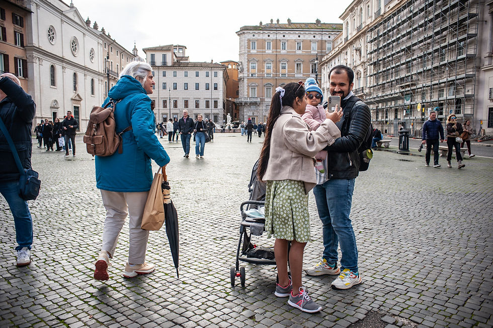Famiglie e turisti a Piazza San Lorenzo in Lucina a Roma durante il Giubileo 2025. Una coppia con una bambina in braccio scatta una foto con lo smartphone, mentre una donna con giacca azzurra e zaino cammina tra la folla. Sullo sfondo, edifici storici e impalcature mostrano il contrasto tra passato e modernità nella città eterna.