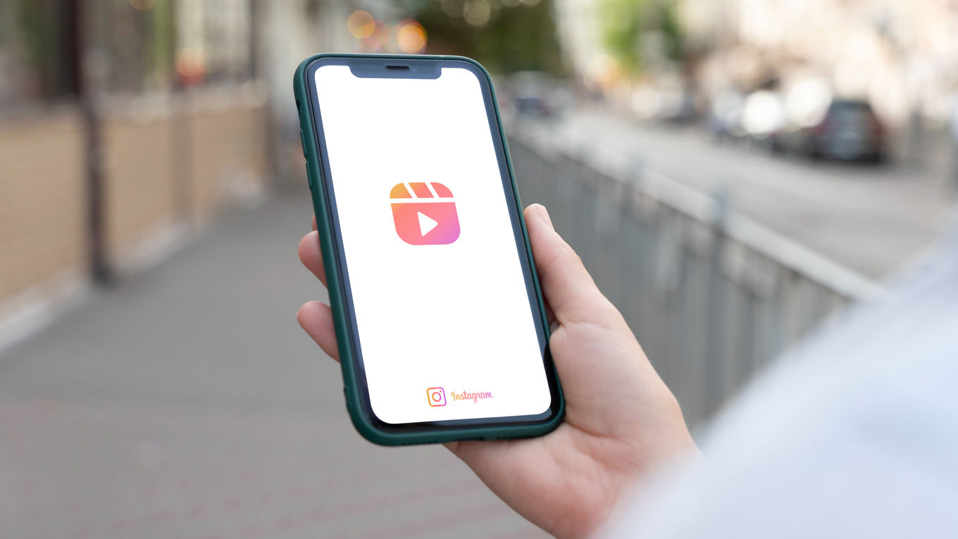 Hand holding a smartphone displaying Instagram Reels logo, with blurred urban street background; phone in a green case, bright screen.