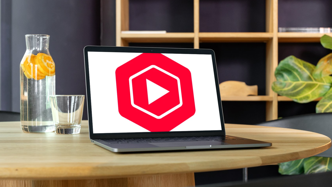Laptop on wooden table showing red play-logo, beside glass pitcher with orange slices and empty glass in a modern room. Youtube studio