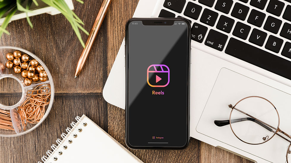 Smartphone sulla scrivania con logo e testo di Instagram Reels, circondato da tastiera, occhiali, quaderno, penna e clip di rame.
