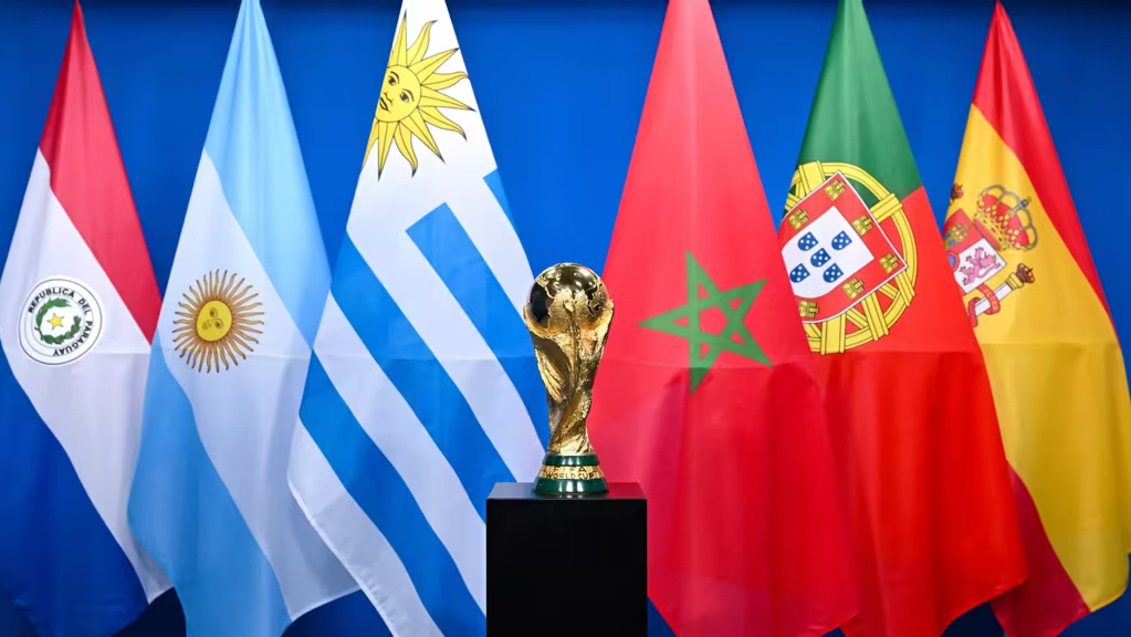 Il trofeo della Coppa del Mondo è posizionato al centro, davanti alle bandiere di Paraguay, Argentina, Uruguay, Marocco, Portogallo e Spagna su uno sfondo blu.