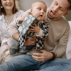 Vater hält Baby am Schoß, das ein Geschenk in der Hand hält, Mutter im Hintergrund