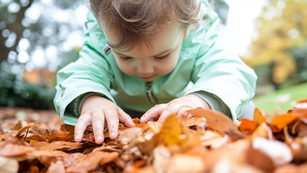 A preschooler in a soft mint green jacket crouches in a pile of wet autumn leaves, pressing small hands into the orange and brown leaf litter in an Australian childcare centre garden