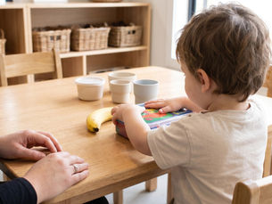 Toddler pressing a colourful lunchbox closed on a kitchen table with a banana and small containers nearby