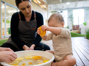 Baby hands squeezing a halved orange over a shallow tub of warm citrus water