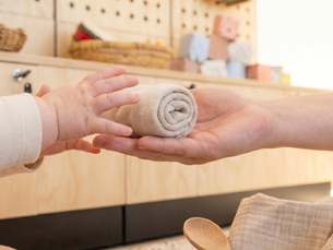 A educators hand offering a rolled sock to a baby's open hands beside a basket of soft household toys on a room floor.