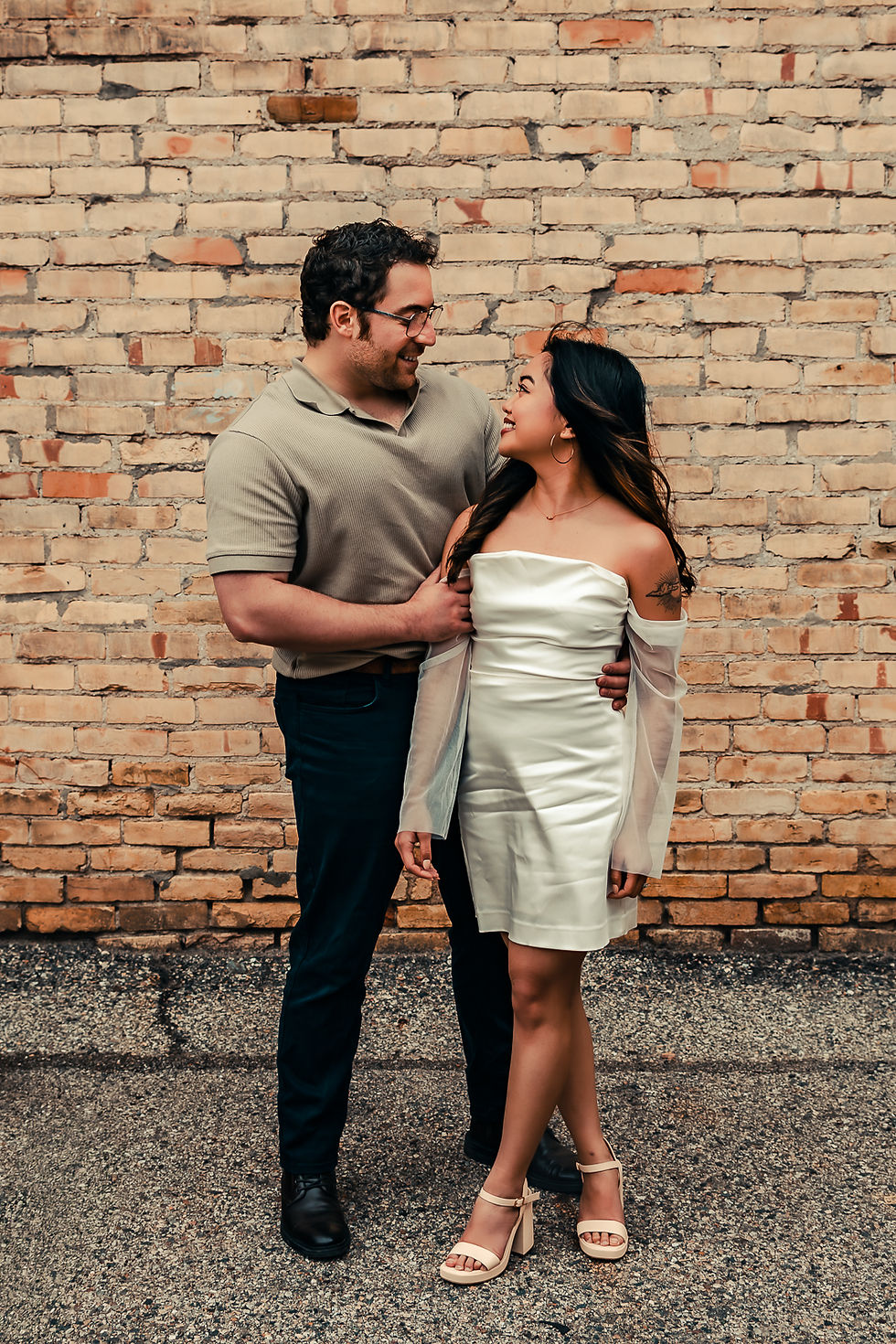 An engaged couple smiles at each other as they stand side by side in front of a brick wall