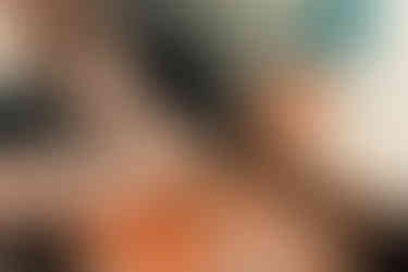 A woman uses both hands and a knife to cut herbs on a wooden cutting board. An open cookbook sits on a stand nearby.