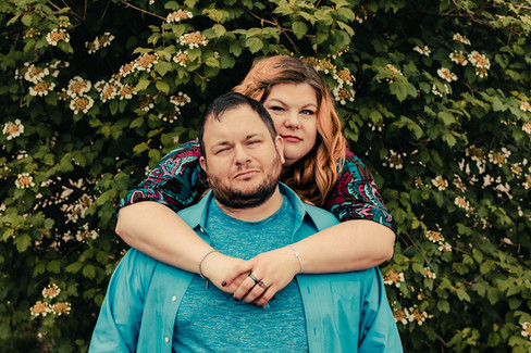 A wife wraps her arms around her husband's neck while both gaze into the camera