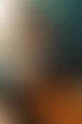 A couple lovingly embraces in front of a wooden kitchen table. One person works to open a cottle of red wine, a clear wine glass next to her. The other kisses her on the neck.