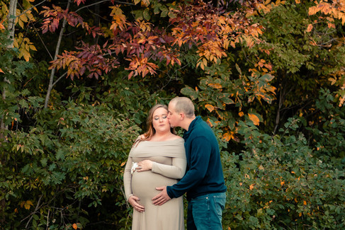 A husband kisses his pregnant wive's cheek while standing in front of a wall of foliage