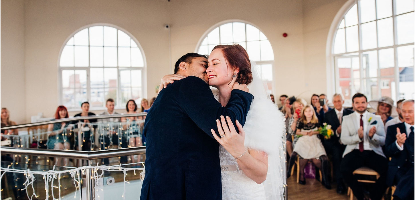 Groom kisses his bride with wedding guests clapping in the background, taking place in the Atrium at the Dome